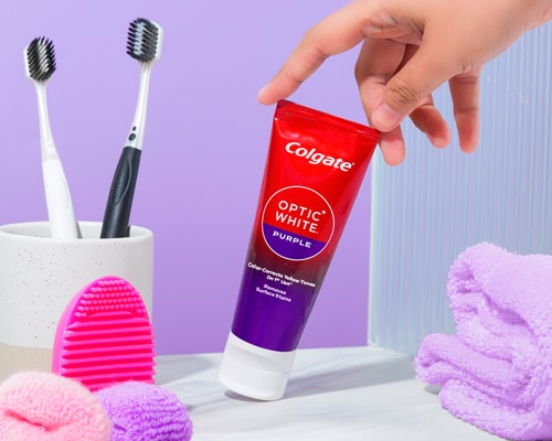 A woman taking the Colgate Optic White Purple Whitening Toothpaste from the slab with toothbrushes in a cup placed on one side and a towel placed on the other side