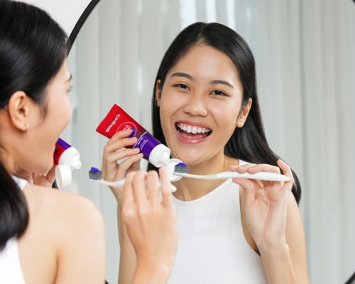 A young smiling woman looking at the mirror and squirting some toothpaste from the Colgate Optic White Purple Whitening Toothpaste tube on to her brush 