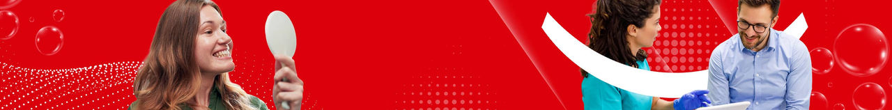 Woman looking in a hand mirror next to a dentist showing a tablet to a male patient against a red background.